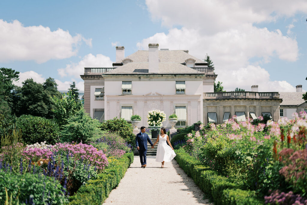Couples photoshoot outfits white midi dress and navy suit walking through colorful cottage garden at Nemours Estate Wilmington Delaware summer engagement photos