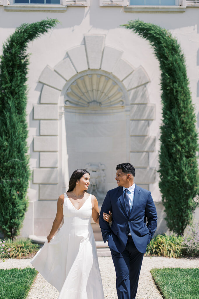 Engagement photo outfit ideas white v-neck midi dress with cross necklace and navy suit walking through Nemours Estate cypress arch garden Delaware