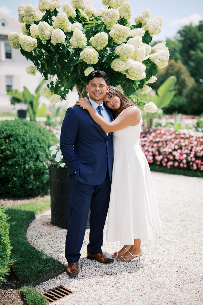 Engagement photo outfits summer white midi dress with strappy silver heels and navy suit couple portrait under white hydrangea tree at Nemours Estate Delaware