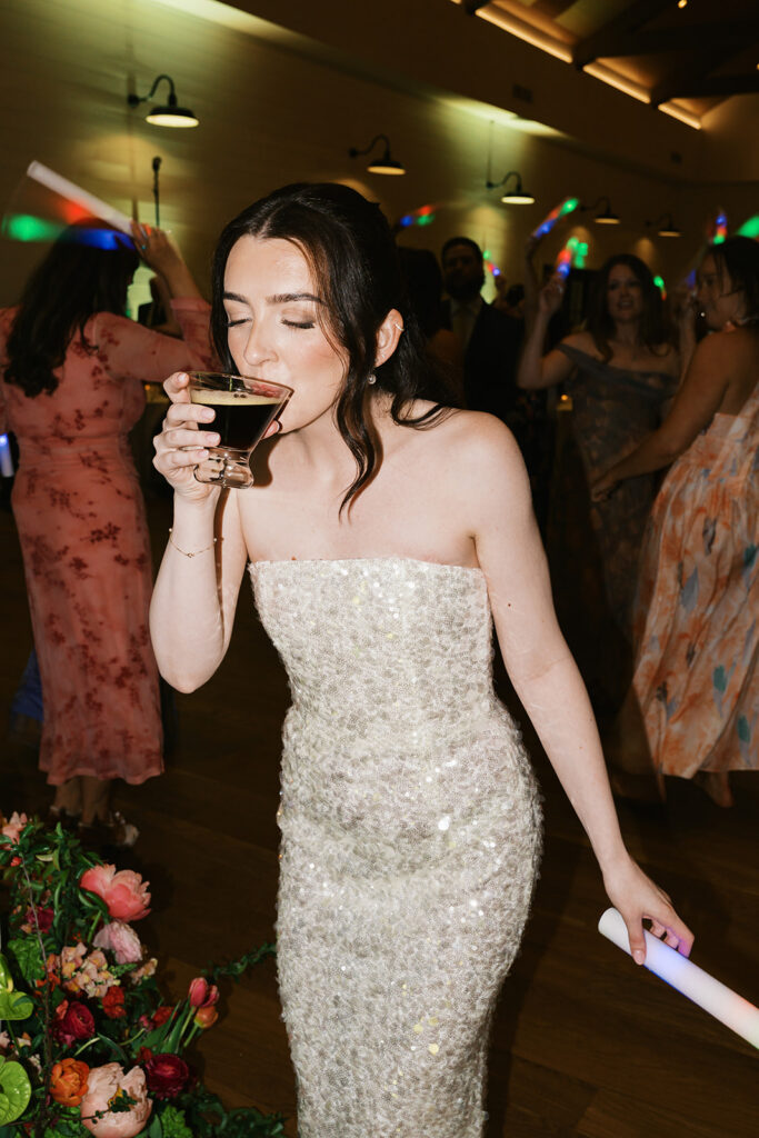 Bride in her sequined second dress sipping an espresso martini on the dance floor as guests dance with glow sticks
