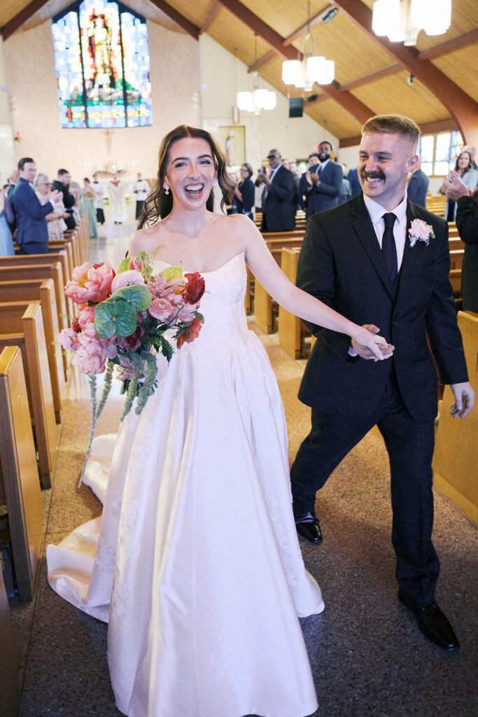 Bride and groom laughing during their recessional up the church aisle, the bride holding a coral peony and green anthurium bouquet