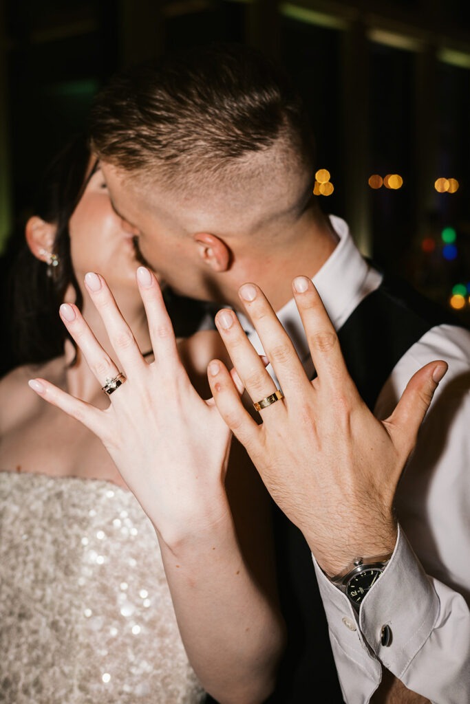 Bride and groom kiss and show off their wedding rings at their reception, with warm bokeh lights behind them