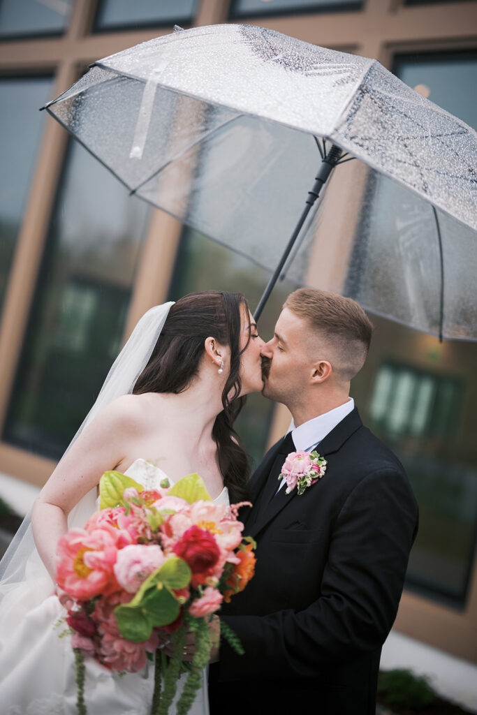 Bride and groom kiss under a clear umbrella in the rain, holding a pink and green bouquet, at a Westwynd Gardens wedding in Honey Brook, PA