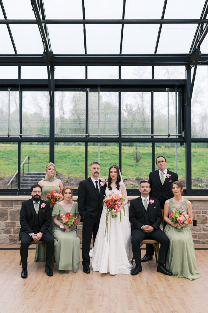 Bride, groom, and sage green bridal party inside the greenhouse with rain on the glass at a Westwynd Gardens wedding in Honey Brook, PA