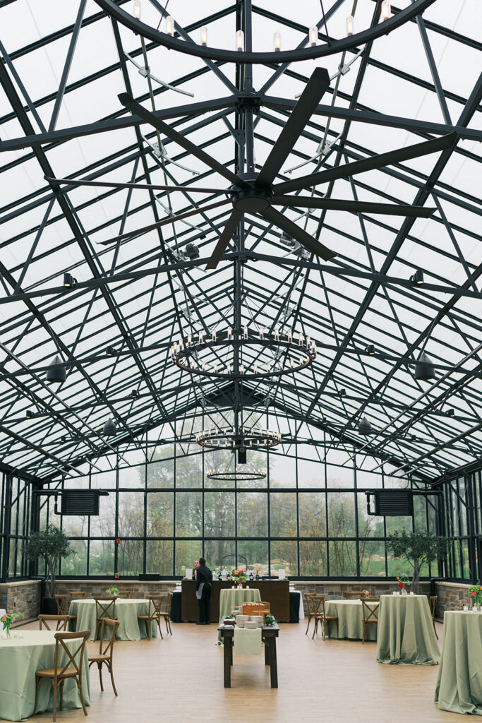 Inside the glass greenhouse at Westwynd Gardens set for a rainy-day cocktail hour, with green linens and a steel-truss ceiling in Honey Brook, PA