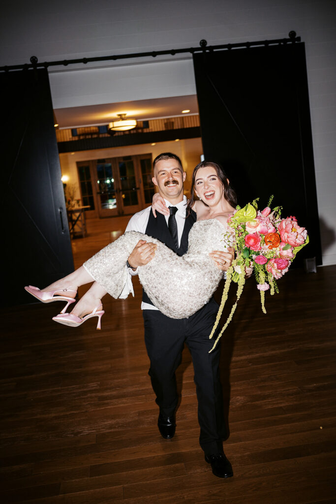 Groom carrying his laughing bride in her sequined second dress through the black barn doors at a Westwynd Gardens reception