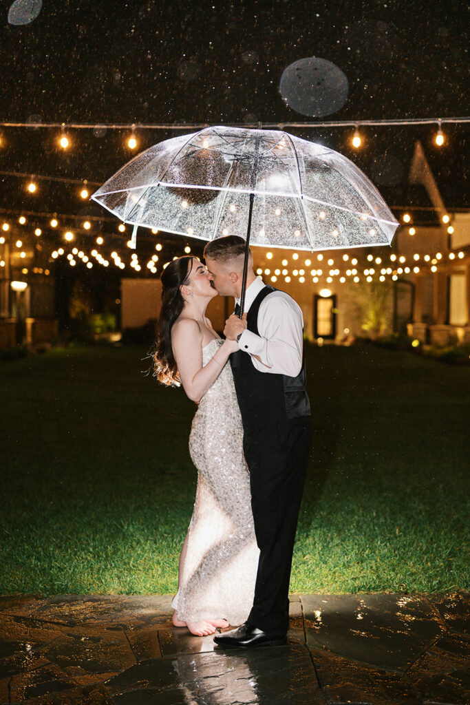 Newlyweds kiss under a clear umbrella in the rain at night beneath glowing string lights, the bride in her sequined second dress, at Westwynd Gardens