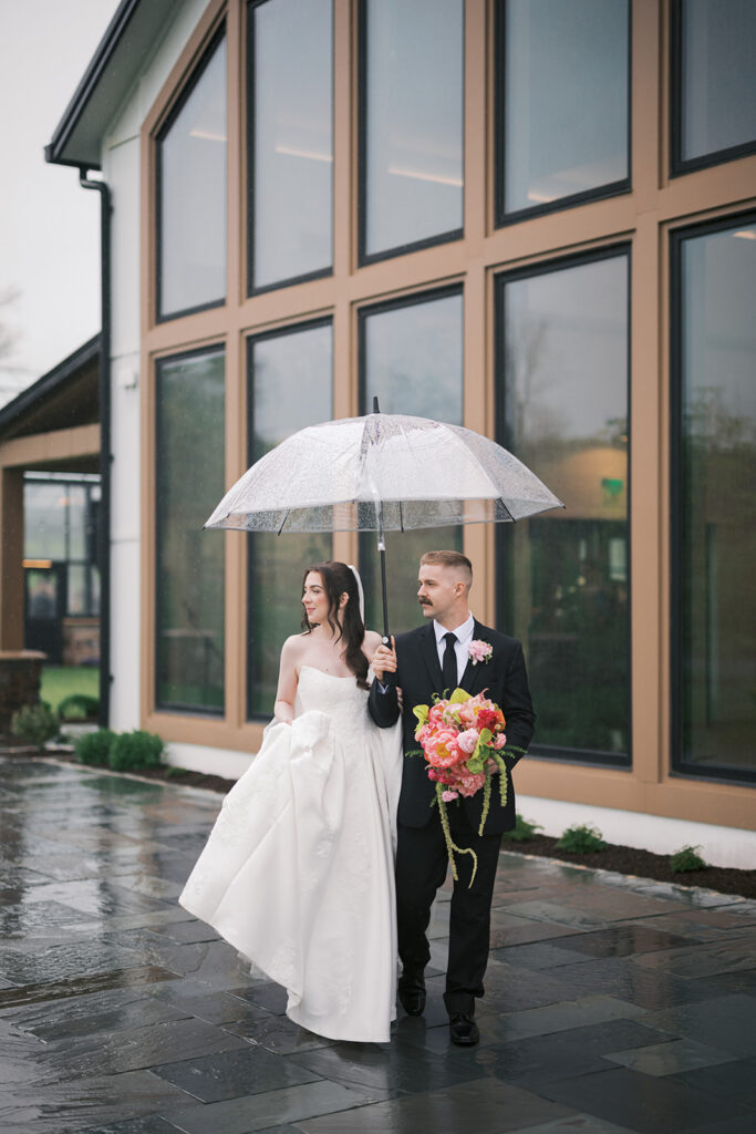 Bride and groom under a clear umbrella in the rain outside the glass venue at a Westwynd Gardens wedding in Honey Brook, PA