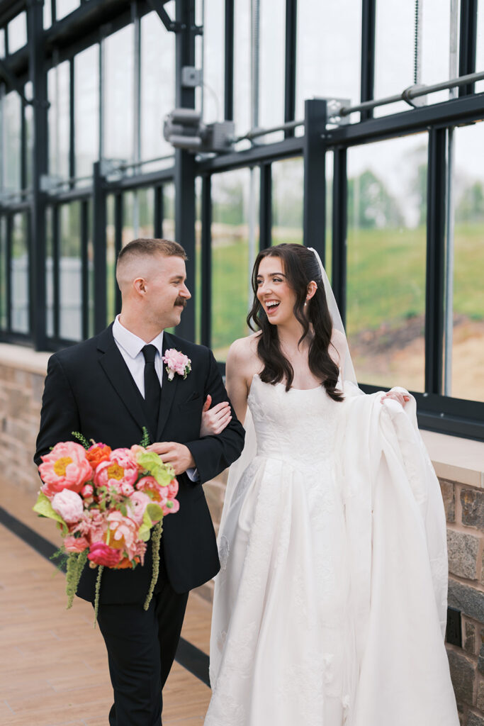 Bride laughing with her groom beside the glass greenhouse, holding a pink and green tropical bouquet at Westwynd Gardens