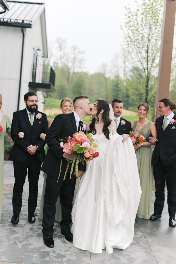 Newlyweds kiss surrounded by their sage green bridal party at a Westwynd Gardens wedding in Honey Brook, PA