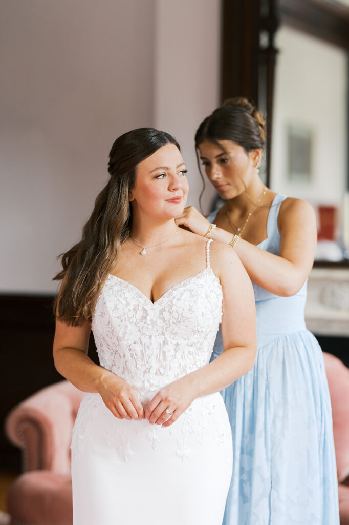 Bridesmaid in a light blue dress helps bride in a corseted white dress clasp her silver pendant necklace