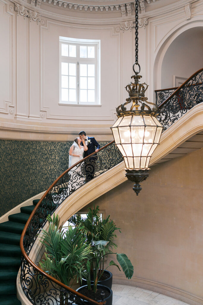 Bride and groom kiss on a velvet green staircase with a detailed wrought iron banister