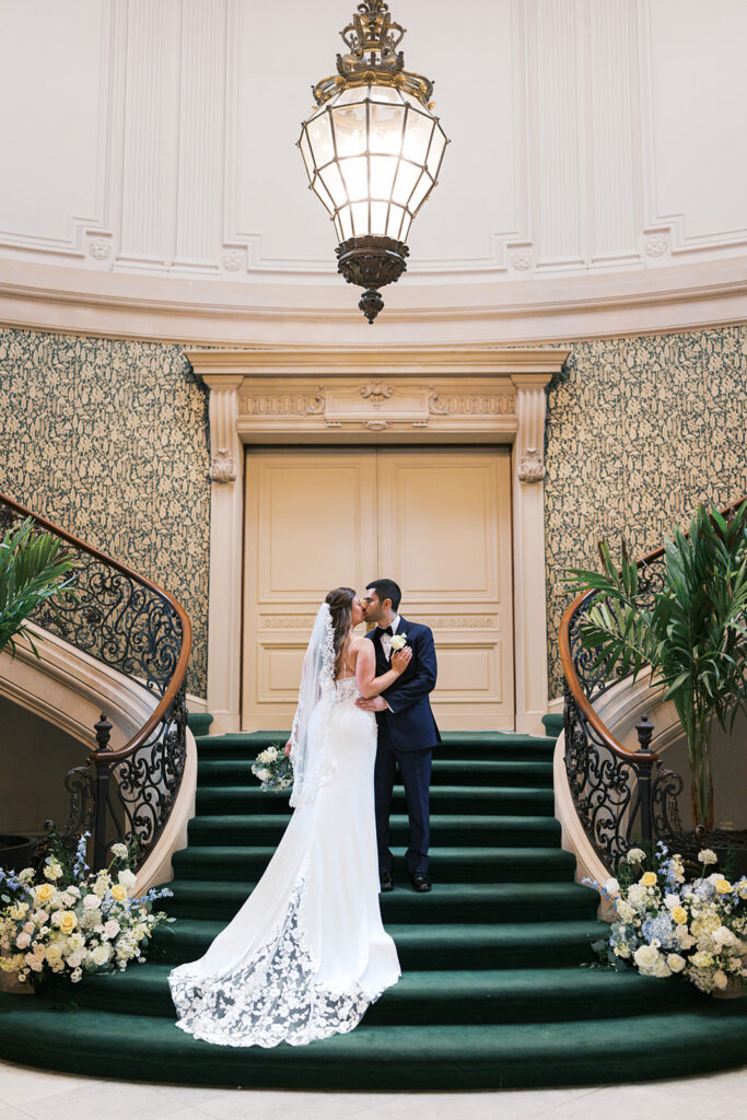 Bride and groom kiss at the base of a grand green staircase at Elkins Estate wedding venue in Philadelphia, photographed by Lindsey Ford