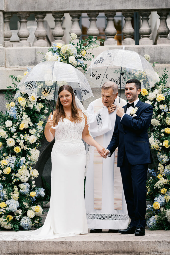 Bride and groom stand with priest and hold clear umbrellas while they smile back at their wedding guests