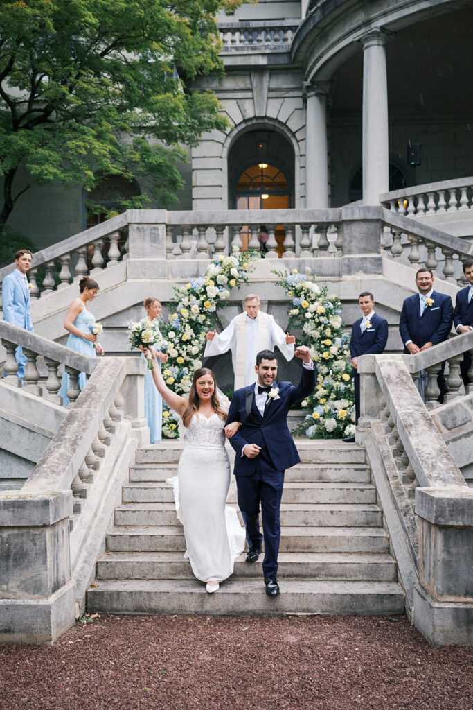 Bride and groom descend a stone staircase with hands raised as their wedding ceremony ends