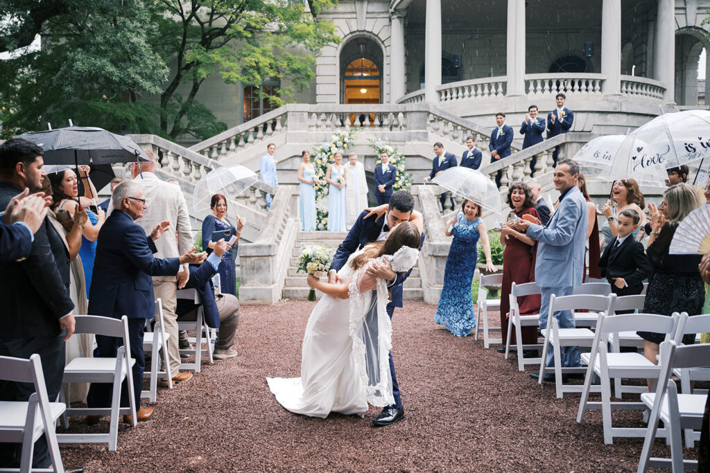 Groom dips bride in the aisle of a rainy outdoor Elkins Estate wedding ceremony while guests holding clear umbrellas look on and cheer