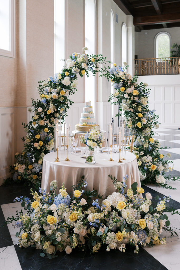 Blue and yellow floral arch over a sweetheart table and tiered cake at an Elkins Estate wedding reception