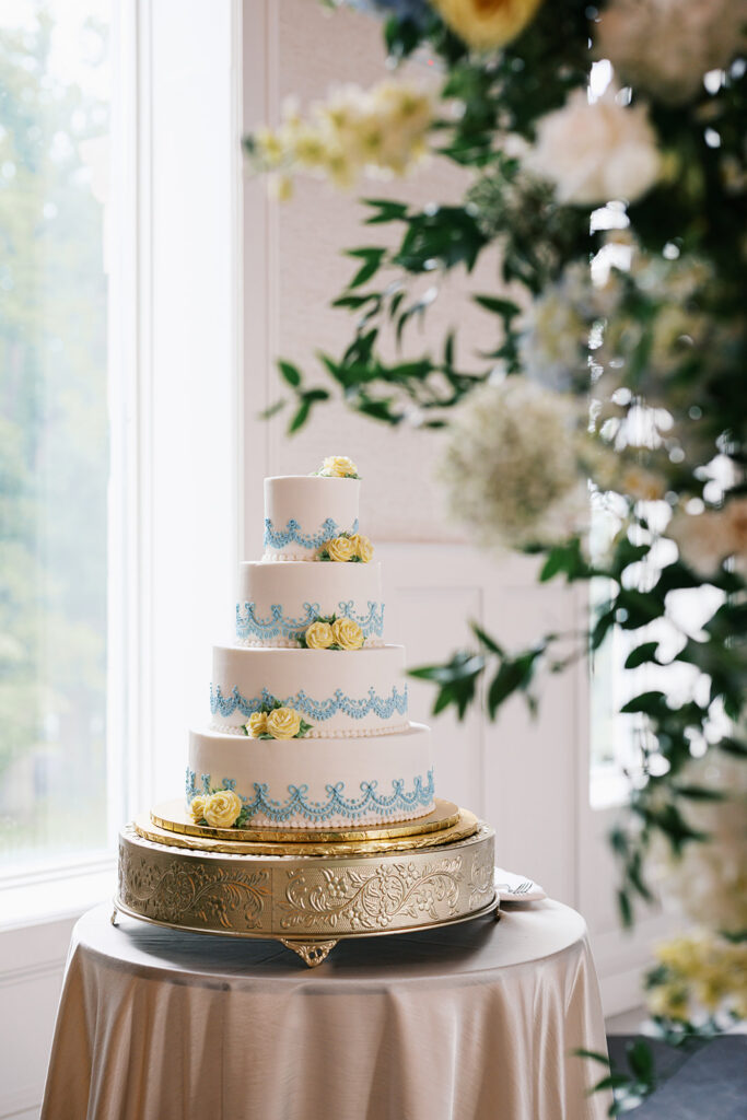 Four tiered white wedding cake with ornate blue piping and yellow rosettes sits on a gold carved cake stand