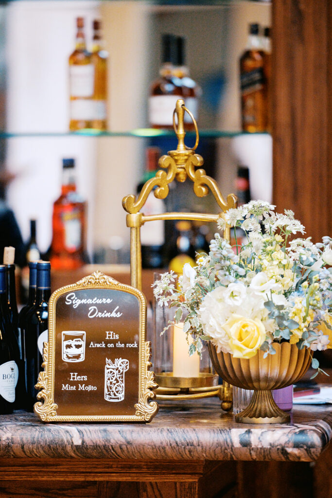 Small mirrored sign listing signature drinks for a wedding sits next to a gold vase holding blue and yellow florals