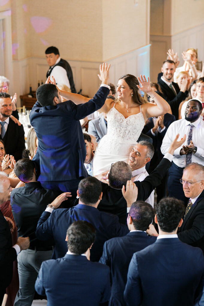 Wedding guests raise bride and groom up on their shoulders as they dance among their wedding guests