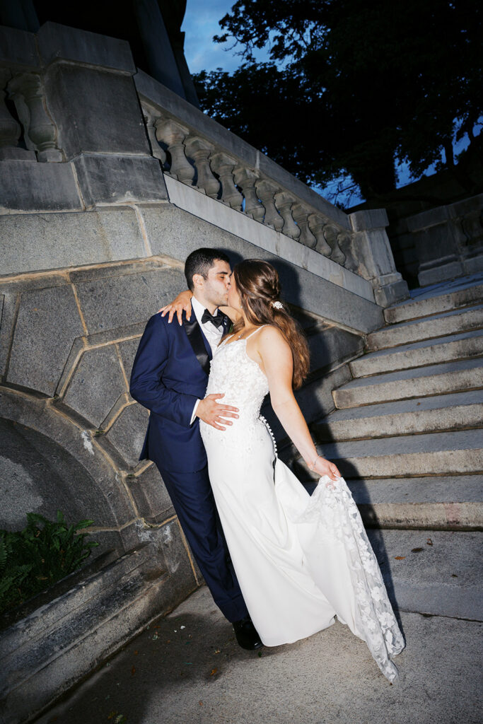 Nighttime flash photography portrait of bride and groom kissing at the bottom of a stone staircase at dusk