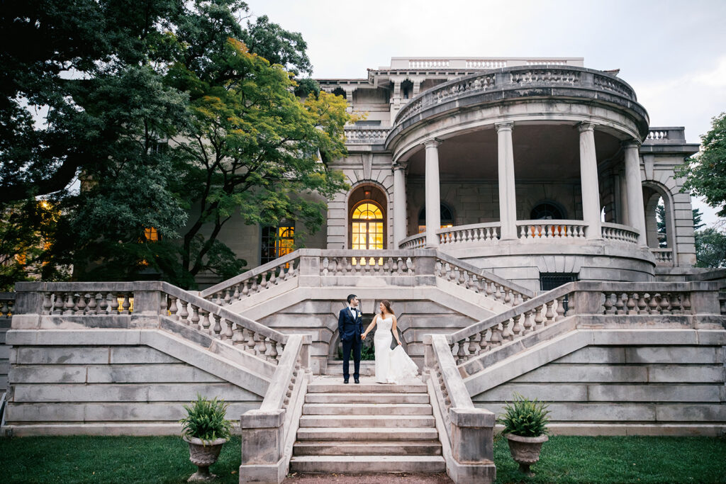 Bride and groom hold hands as they stand at the top of a grand staircase outdoors at Elkins Estate wedding venue in Philadelphia