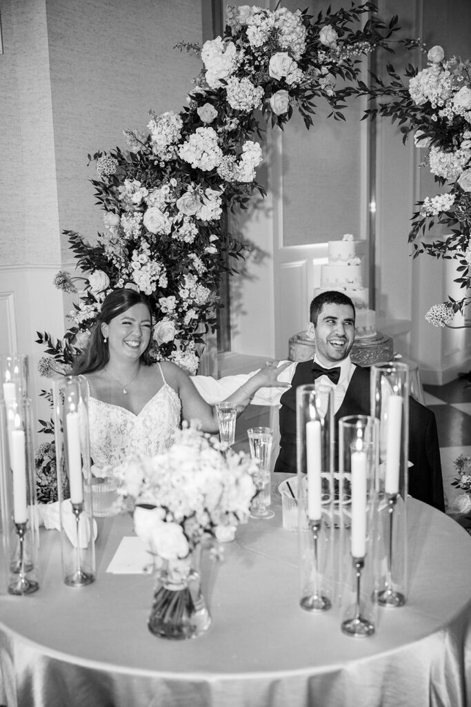 Bride and groom sit together laughing at their sweetheart table under a floral arch