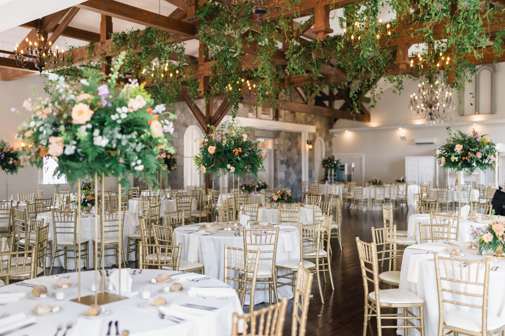 Round tables are decorated for a wedding reception under wooden beams hung with greenery inside the Loch Aerie Mansion reception space