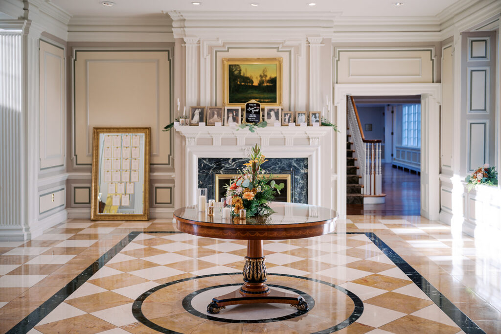 Circular wooden table decorated with autumn florals sits in the lobby of Radnor Valley Country Club in front of a marble mantle and a mirrored seating chart